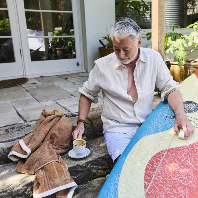 Elderly man sitting on a porch with a coffee cup next to him, while he is holding a surf board.